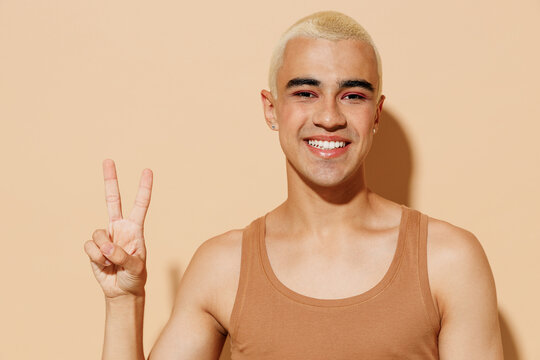 Young trendy fun cool friendly fun blond latin gay man 20s with make up in beige tank shirt showing victory sign isolated on plain light ocher background studio portrait People lgbt lifestyle concept.