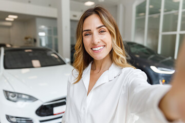 Close up woman customer female buyer client in white shirt doing selfie shot pov on mobile phone choose auto want to buy new automobile in car showroom vehicle salon dealership store motor show inside