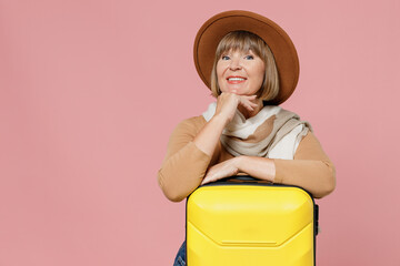 Traveler tourist fun mature elderly senior lady woman 55 years old wear brown shirt hat scarf hold suitcase bag put hand prop up on chin isolated on plain pastel light pink background studio portrait.