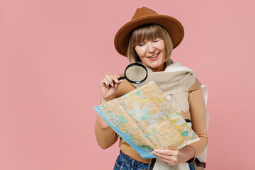Traveler tourist smiling mature elderly senior lady woman 55 years old wear brown shirt hat scarf hold examine map with magnifying glass isolated on plain pastel light pink background studio portrait