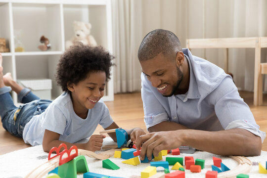 Happy young african american father playing wooden toys with cute little child son lying on floor carpet, having fun enjoying weekend leisure activity together at home, single parenting concept.