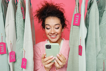 Young satisfied smiling female costumer woman wear sweater stand near clothes rack with tag sale in store showroom hold in hand use mobile cell phone isolated on plain pink background studio portrait.