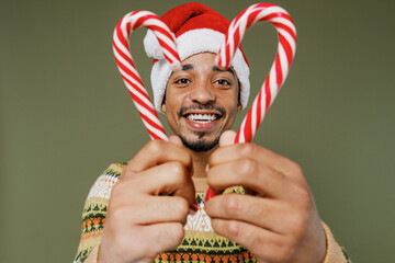 Young fun african man in sweater red Christmas Santa Claus hat hold in hand two candy cane lollipops isolated on plain green khaki background studio portrait. Happy New Year 2022 celebration concept.