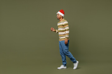 Fototapeta na wymiar Full size side view young african man 20s in knitted sweater red Christmas Santa Claus hat walking go isolated on plain green khaki background studio portrait. Happy New Year 2022 celebration concept.