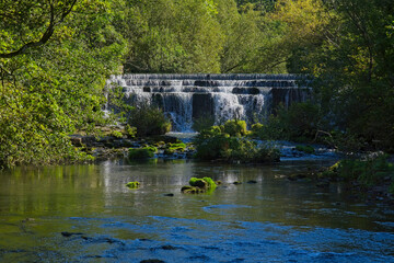 Fototapeta na wymiar The River Wye tumbles over Monsal Dale weir surrounded in trees and bushes.