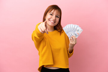 Redhead girl taking a lot of money isolated on pink background pointing front with happy expression © luismolinero