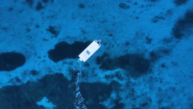 High view of a boat slowly moving across a reef structure in dark blue water. Drone view