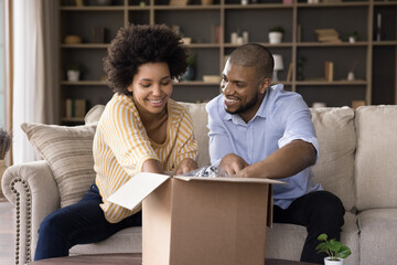 Happy curious young african american family couple opening big carton box, enjoying unpacking...