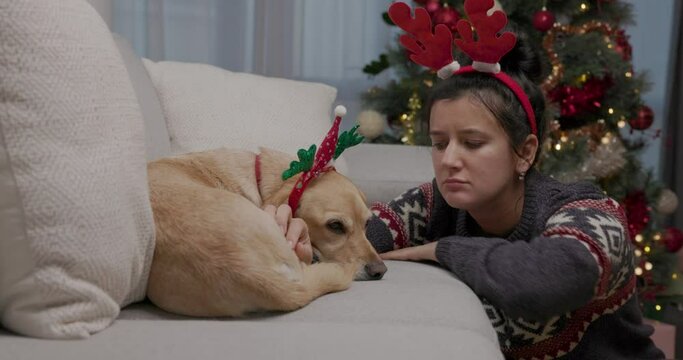 Sad lonely young woman sitting frustrated next to her yellow dog by couch at home during festive season, wearing Christmas antlers