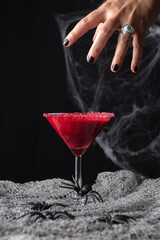 View of woman's hand over glass of red cherry cocktail for Halloween on table with spiders, black background, vertical