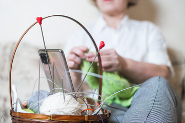 Faceless old woman knits and watches online training on a smartphone. Close-up of female hands with yarn and knitting needles.