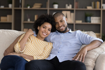 Obraz premium Smiling bonding mixed race young married couple homeowners tenants looking at camera, relaxing on comfortable sofa on weekend at home, spending leisure weekend time together, family portrait.