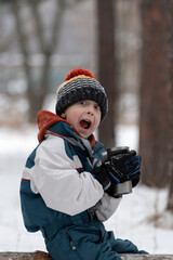 Fototapeta na wymiar Cheerful boy in knitted hat with thermos in his hands on snow-covered city park background. Picnic in the winter forest. Open mouth wonder