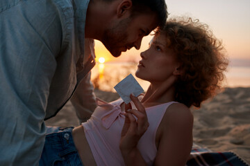 Beautiful young couple ready for making love on the beach at sunset. Woman is showing a condom to...