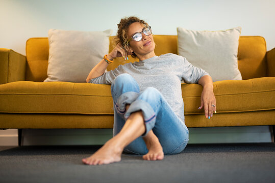 Relaxed mature woman sitting on the floor barefoot at home looking outside with yellow sofa in background. Happy female people young adult enjoy apartment leisure activity relaxing and resting