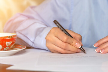 Young woman with a beautiful manicure in a blue shirt writes notes with a pen on paper sheets.