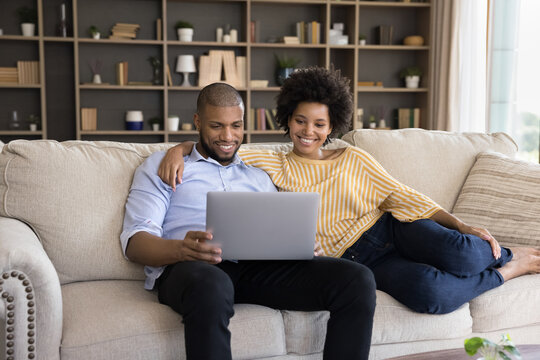 Smiling Loving Bonding Young Mixed Race Family Couple Using Computer, Watching Comedian Movie, Buying Goods In Internet Store, Booking Tickets Or Hotel, Reading News Or Web Surfing Useful Information.