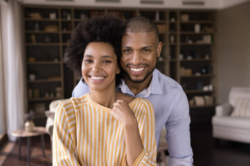 Portrait of happy loving young mixed race family couple posing in modern living room. Smiling bonding millennial homeowners celebrating moving into own apartment, good family relations concept.