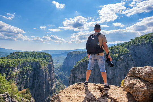 Full body back view of unrecognizable male hiker with backpack and photo camera in hand standing on edge of rocky cliff in highlands
