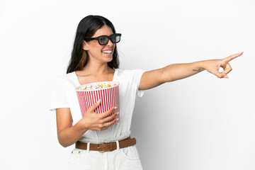 Young caucasian woman isolated on white background with 3d glasses and holding a big bucket of popcorns while pointing away