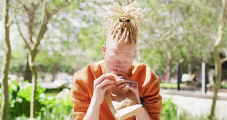Albino african american man with dreadlocks sitting in park with bike eating sandwich - Powered by Adobe