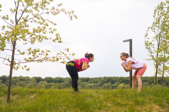 Wide shot of cheerful fitness female trainer giving personal training to motivated overweight young woman outdoor in summer day. Instructor help fat woman lose weight outside doing squats outside.