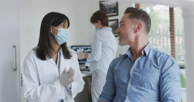 Biracial female dentist with face mask preparing male patient at modern dental clinic