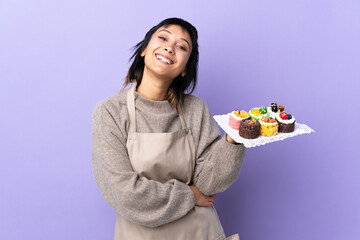 Young Uruguayan woman holding lots of different mini cakes over isolated purple background laughing