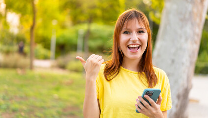 Fototapeta premium Young pretty redhead woman at outdoors using mobile phone and pointing to the lateral