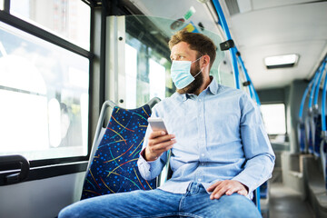 Young man on a bus rides to his destination, wearing a face mask for virus protection, uses a...