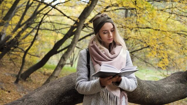 Female Adult Girl Standing in Autumn Forest with Yellow Leaves Reading a Book on Nature. Pretty Woman Rest and Relax with a book in hands. Blue Eyes Blonde is Studying Outdoors. Inspiration Motivation