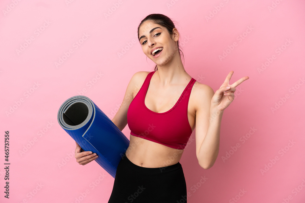 Poster Young sport caucasian woman going to yoga classes while holding a mat smiling and showing victory sign