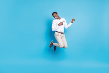 Photo of positive funky dark skin man wear white shirt smiling jumping high pointing empty space isolated blue color background