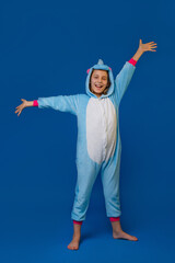 A girl in a cozy blue unicorn costume in full height, with her arms outstretched, poses in the studio on a blue isolated background. Place for text