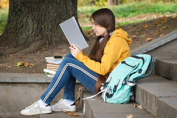 A girl of 13 years old sits on the stairs and reads a book in the park, goes to school exams.