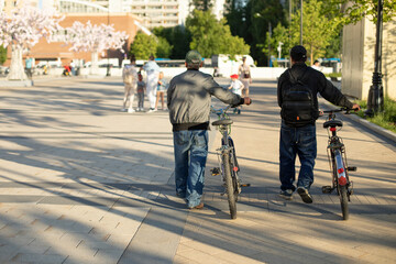 Two people with bicycles walk around the square of the city. Men in jeans and gray jackets.