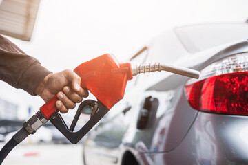 A gas station worker holds a fuel dispenser to fill the car with fuel. A young man's hand holds a...