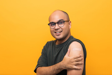 Vaccination campaign. Young handsome indian man getting coronavirus vaccine Immune and showing arm with bandage plaster on isolated yellow background.