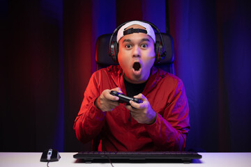 Excited and shocked face of Asian gamer with headphone holding joystick playing video game online sitting on chair at living room. Indian professional gamer streaming on social playing game very fun - obrazy, fototapety, plakaty