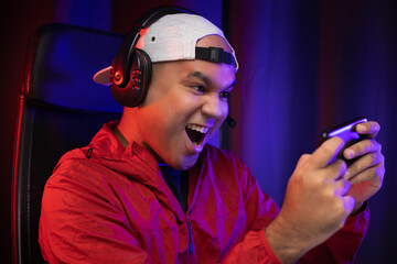 Playing video game on smartphone. Young asian handsome man sitting on chair holding cellphone in living room. Happiness Streamer Indian man wearing headset playing game online in the darkroom.