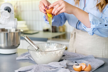 Close up of dough preparation process. Hands break an egg into a bowl of flour in a light kitchen in light clothes