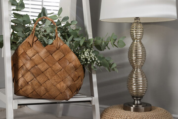 Stylish straw bag with beautiful bouquet and lamp indoors