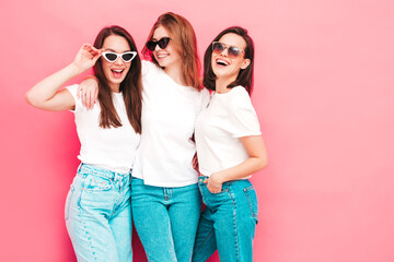 Three young beautiful smiling hipster female in trendy same summer white t-shirt and jeans clothes. Sexy carefree women posing near pink wall in studio. Positive models