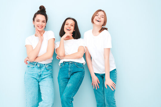 Three young beautiful smiling hipster female in trendy same summer white t-shirt and jeans clothes. Sexy carefree women posing near light blue wall in studio. Cheerful and positive models having fun