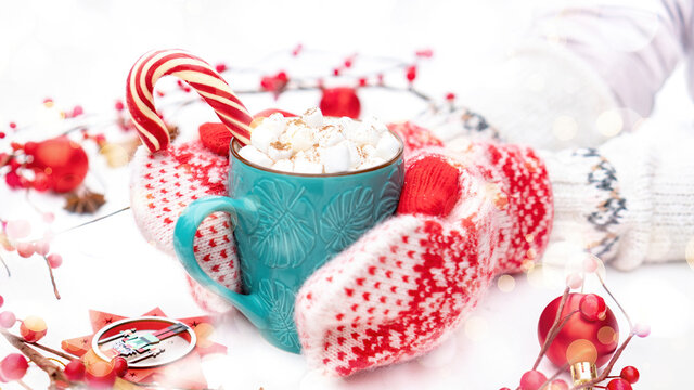 Hands in knitted woolen mittens hold a mug of hot cocoa with marshmallows and a candy cane. A traditional Christmas photo in red and white colors with a blurred white background with bokeh.