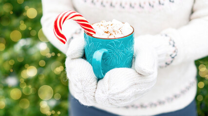 A girl in a white knitted sweater and mittens holds out a mug with a delicious Christmas drink. A cup of hot cocoa with marshmallows and candy cane in hands close up. Giving gifts for Christmas.