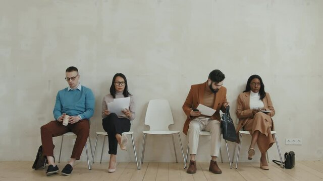 Timelapse medium shot of diverse group of men and women in formal wear entering frame and sitting down on chairs to wait for job interview