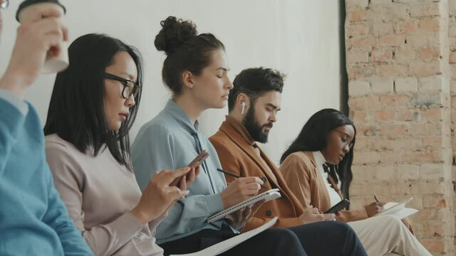 Side view shot of diverse group of job applicants in formal wear sitting in line and waiting for interview. They are drinking coffee, making notes and using mobile phones