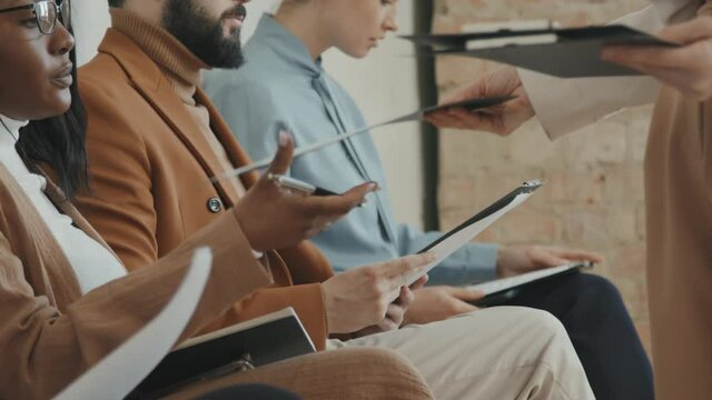 Side view close up shot of unrecognizable HR manager giving clipboard with application forms to people in formal wear sitting in row and waiting for job interview
