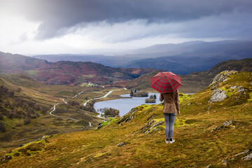 Fototapeta premium A tourist woman with a scotish pattern umbrella enjoys the view over the highlands to Lock Knokie in Scotland during autumn time